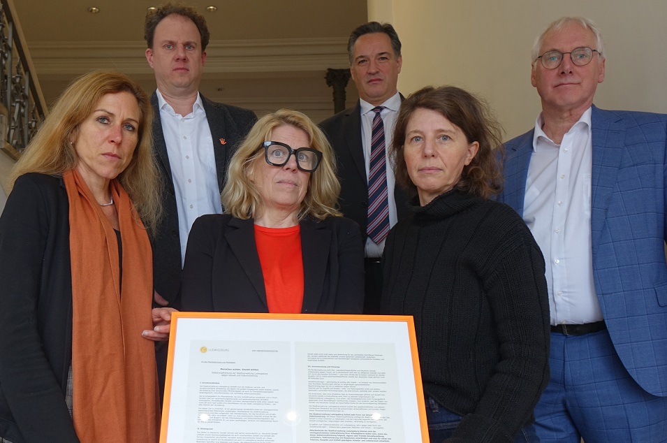 Sechs Personen (vorne drei Frauen, dahinter drei Männer) stehen auf einer Treppe und blicken ernst in die Kamera. Vor ihnen steht ein Ausdruck der Selbstverpflichtung der Stadtverwaltung gegen Gewalt und Diskriminierung in einem orangenen Rahmen.