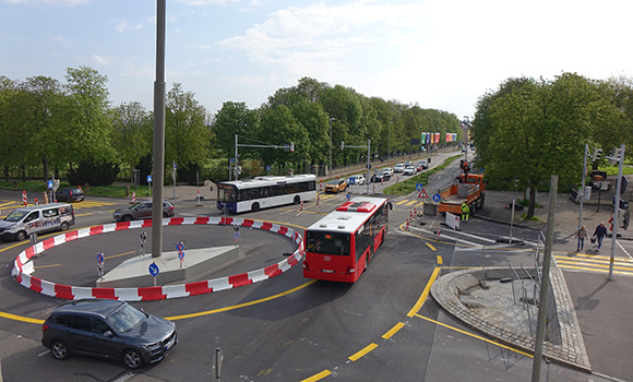 Im Kreisverkehr fahren Autos und Busse. Menschen gehen über einen Zebrastreifen.