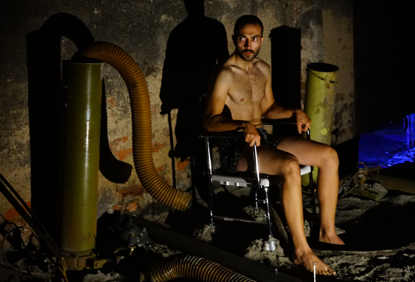 Ein junger Mann sitzt halb nackt im dunkeln auf einem Stuhl.