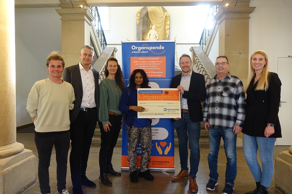 sieben Personen vor einem Treppenaufgang, hinter ihnen ein Organspende-Aufsteller. Zwei der Personen halten geneinsam einen symbolischen Organspendeausweis zwischen sich.                 