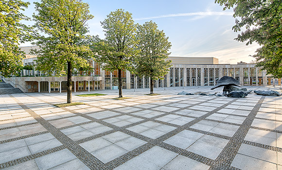 Das Bild zeigt einen großen Platz vor dem Gebäude des Forums am Schlosspark. Auf dem Platz stehen Bäume und ein Brunnen.