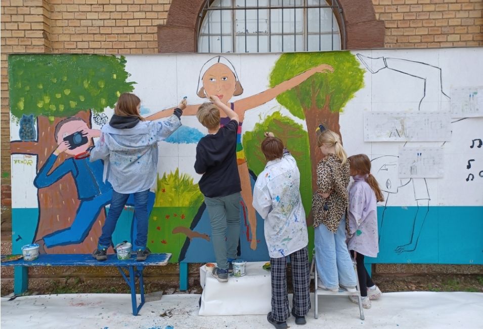 Fünf Kinder stehen in einem Hof und Malen auf einer lebensgroßen Wand. 