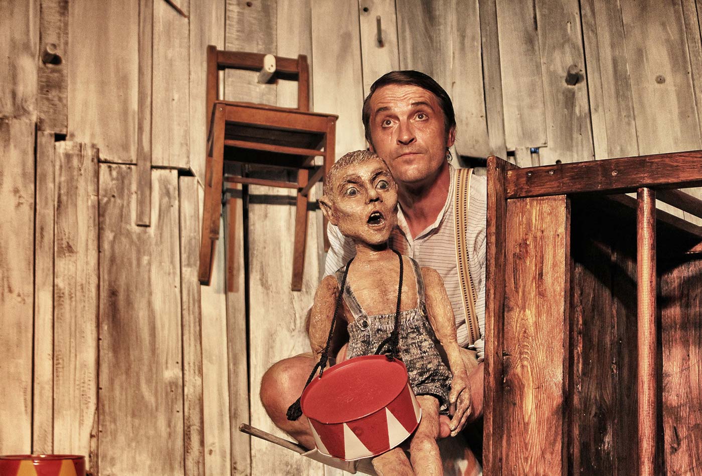 EIn Schauspieler steht mit einer Puppe in der Hand auf eienr Bühne.
