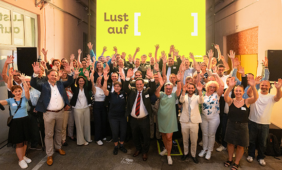 Auf eine große Leinwand ist das Motiv "Lust auf [ ]" projiziert. Davor stehen rund 50 Teilnehmende.