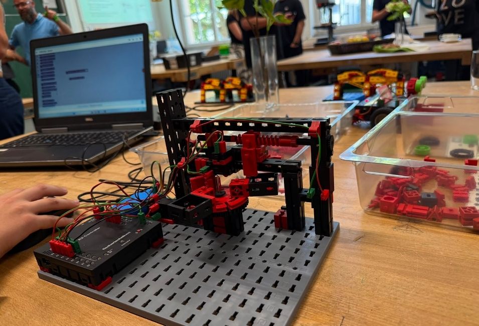 Auf einem Tisch steht ein programmierbares Fischertechnik-Modell mit Kabeln und Steuerung. Daneben befinden sich ein Laptop und weitere Fischertechnik-Baukästen. 