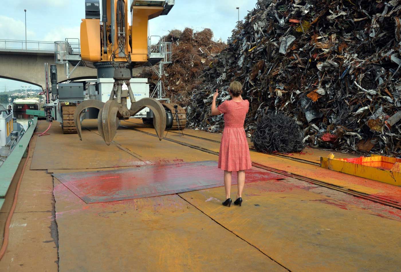 Eine Frau im roten Kleid steht vor einem Greifarm. Daneben Berge von Metallschrott.