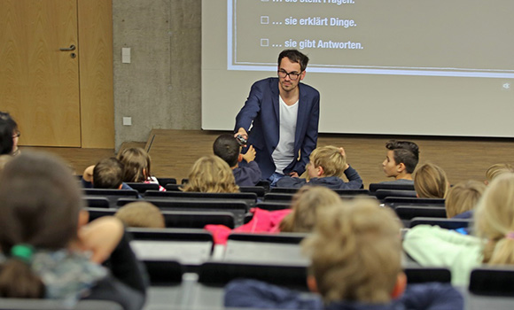 Kinder sitzen in einem Hörsaal. Vor den Kindern steht ein Mann und erklärt ihnen etwas.