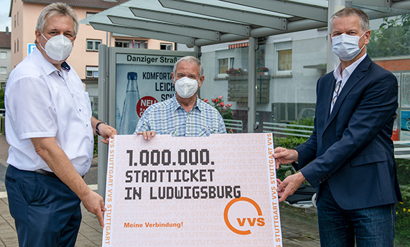 Drei Männer, alle mit Atemschutzmasken, halten ein übergroßes Stadtticket. Dieses hat die Aufschrift: 1.000.000 Stadtticket in Ludwigsburg.Die Männer stehen vor einer Bushaltestelle in der Danziger Straße.