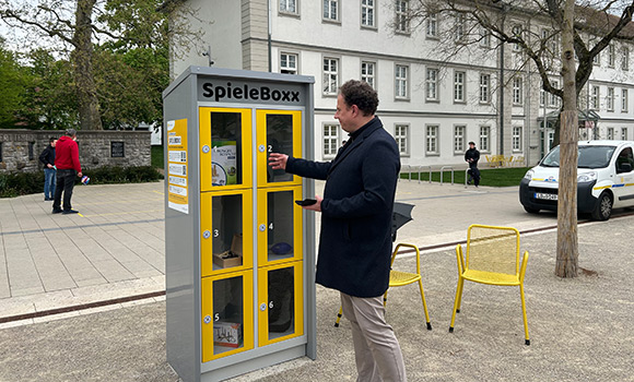 OB Knecht steht vor einer Spielebox. Seine Hand ist am Griff eines der sechs Fächer der Box.