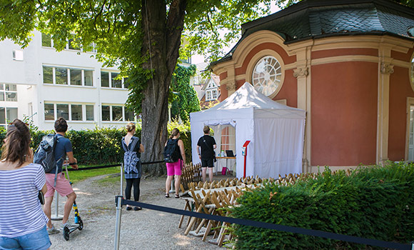 Am Eingang eines kleinen barocken Gebäudes steht ein weißes Zelt. Davor stehen fünf Personen in einer Schlange.